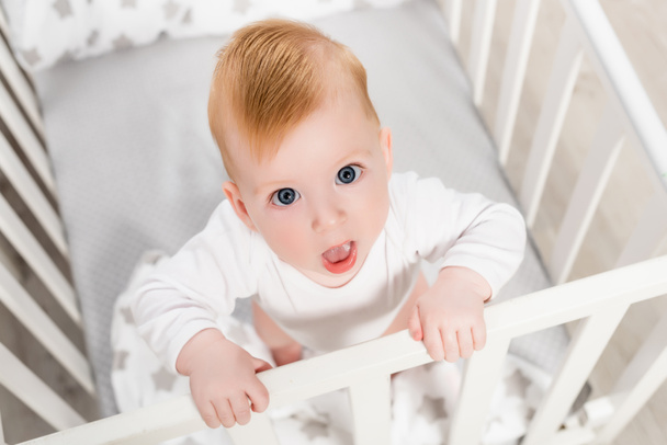 overhead view of infant child looking at camera with open mouth while standing in crib - Fotografie, Obrázek