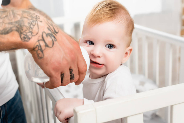 selective focus of tattooed man feeding infant child standing in cot  - Photo, Image