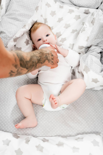 selective focus of tattooed man feeding infant son from baby bottle - Foto, imagen