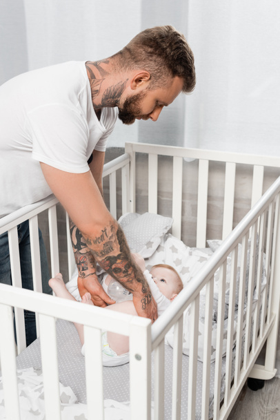 joven hombre tatuado tocando al bebé hijo acostado en la cuna con biberón - Foto, Imagen