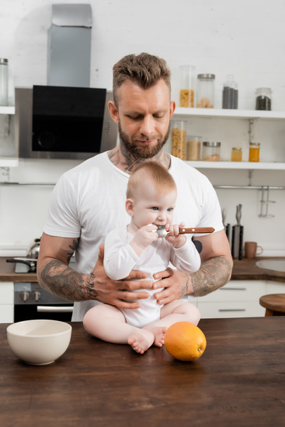 infant child holding spoon near mother while sitting on kitchen table near young father - Photo, Image