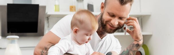 horizontal image of young man talking on smartphone in kitchen during breakfast with infant son - Foto, Bild