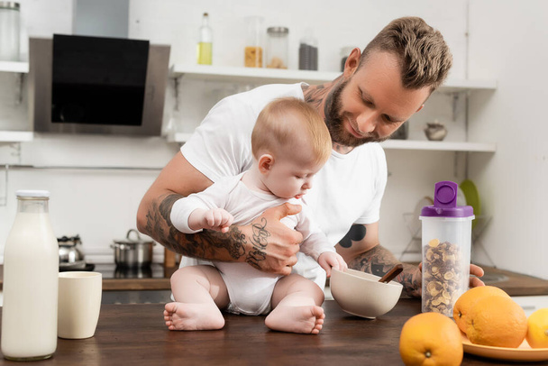 Младенец трогает миску с завтраком, сидя на кухонном столе рядом с молодым татуированным отцом - Фото, изображение
