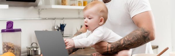 colpo panoramico di bambino neonato che tocca il computer portatile vicino al padre tatuato in cucina - Foto, immagini