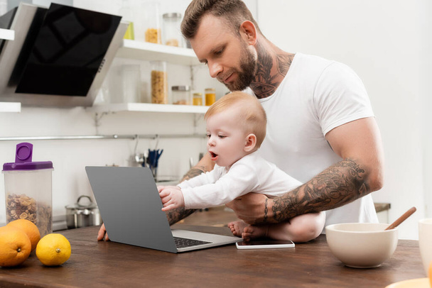 Ο άντρας με τα τατουάζ κρατάει το μωρό γιο του αγγίζοντας το λάπτοπ ενώ κάθεται στο τραπέζι της κουζίνας - Φωτογραφία, εικόνα