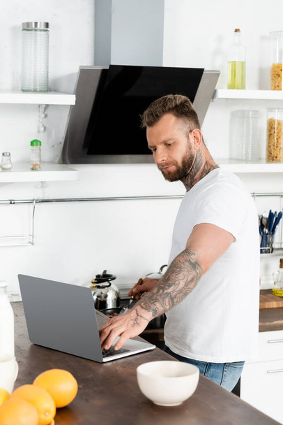 young, bearded freelancer in white t-shirt using laptop near oranges and bowl in kitchen - Fotoğraf, Görsel