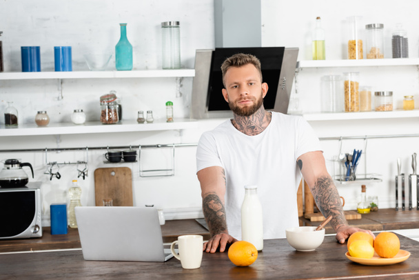 bearded, tattooed blogger in white t-shirt looking at camera near breakfast and laptop in kitchen - Photo, Image