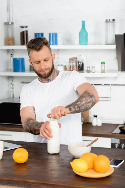 młody brodaty mężczyzna w białej koszulce otwierającej butelkę świeżego mleka podczas śniadania w kuchni - Zdjęcie, obraz