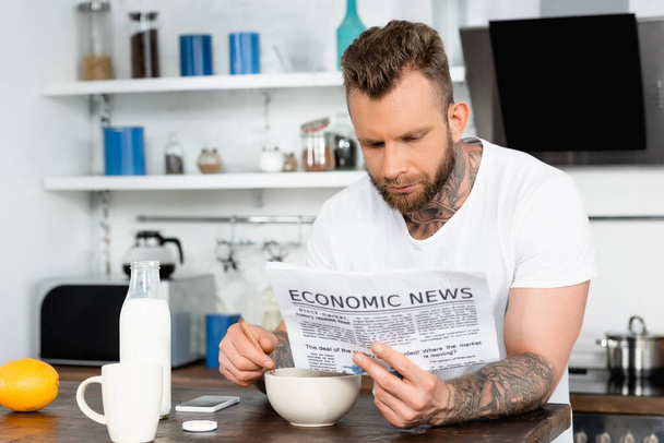 young bearded man in white t-shirt reading newspaper during breakfast in kitchen - Foto, Bild