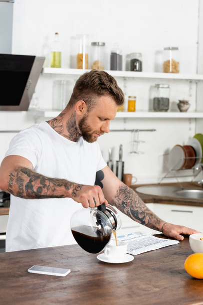 白いTシャツに身を包んだ男が新聞を読み、キッチンテーブルの上に携帯電話の近くにコーヒーを注ぐ - 写真・画像