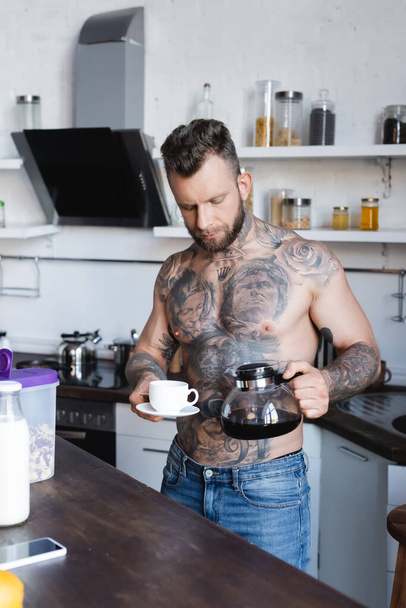 sem camisa, homem tatuado segurando panela de café e xícara na cozinha - Foto, Imagem
