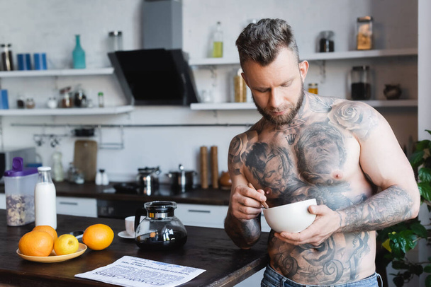 muscular, tattooed man holding bowl with breakfast while standing in kitchen - Foto, Imagem