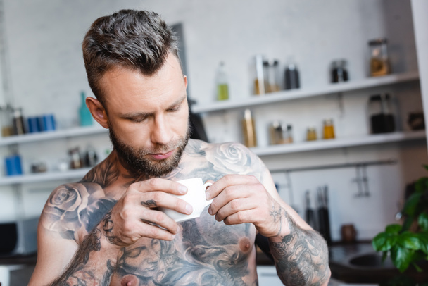 young tattooed man holding cup of coffee in kitchen - Фото, изображение