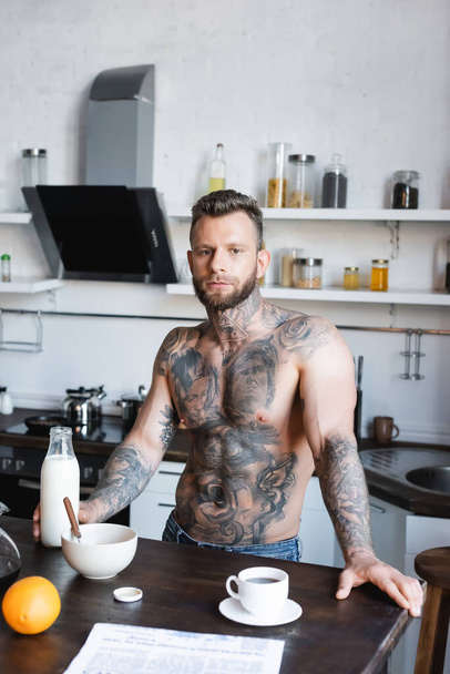 muscular, tattooed man looking at camera while standing in kitchen during breakfast - Foto, imagen