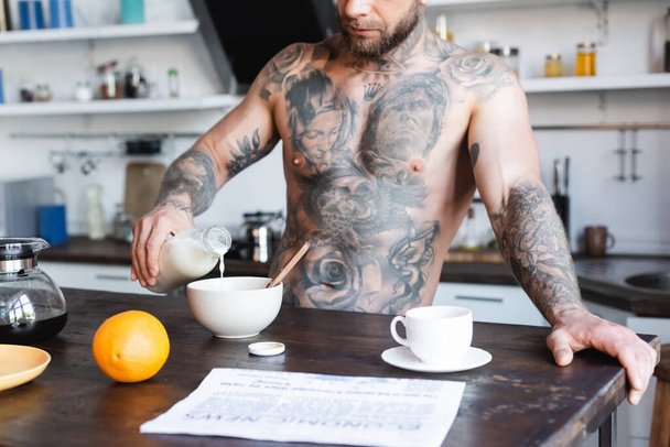 kivágott kilátás tetovált férfi öntés friss tejet tálba reggeli közben a konyhában közelében újság az asztalon - Fotó, kép