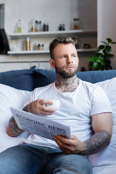 selective focus of bearded tattooed man holding coffee cup and newspaper while sitting in bed - Zdjęcie, obraz