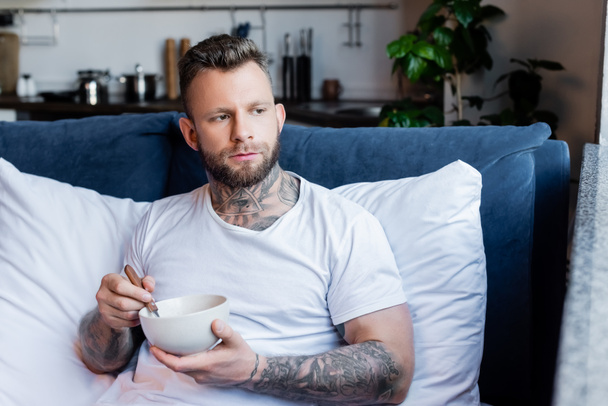young tattooed man looking away while having breakfast in bedroom - Foto, afbeelding