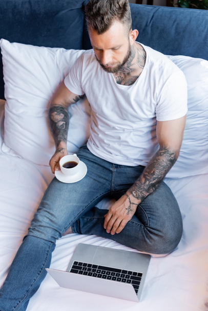 hoge hoek uitzicht van getatoeëerde freelancer in wit t-shirt en jeans met behulp van laptop en met een kopje koffie in bed - Foto, afbeelding