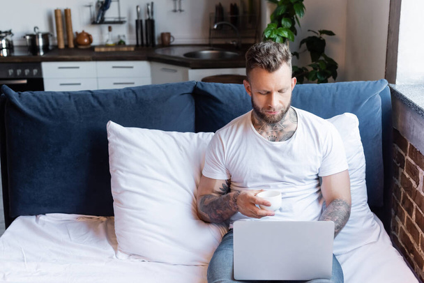 joven, tatuado freelancer utilizando el ordenador portátil en la cama mientras sostiene la taza de café - Foto, Imagen