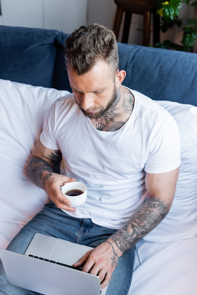 high angle view of tattooed freelancer holding cup of coffee while using laptop in bedroom - Fotó, kép