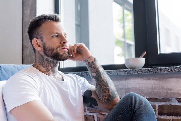 серьезный татуированный мужчина сидит у окна дома и смотрит в сторону - Фото, изображение