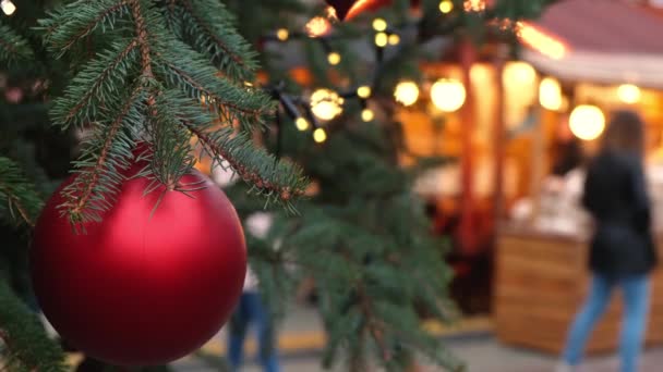 Close-up van een rode kerstbal hangend aan een natuurlijke nieuwjaarsboom op een stadsmarkt. Wazige menigte van mensen lopen op een feestelijk plein. - Video