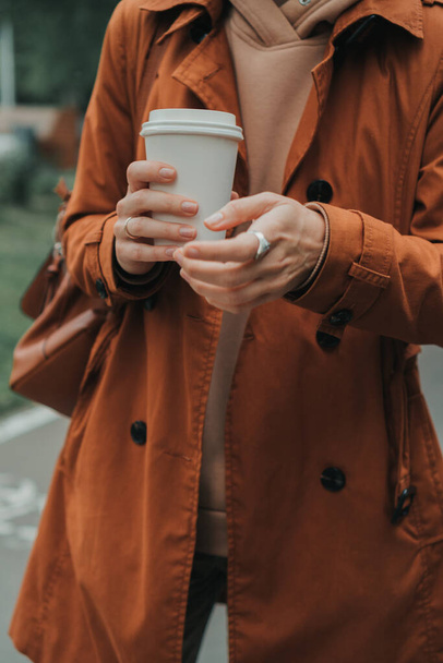 Bir kız parkta turuncu yağmurlukla duruyor ve elinde bir bardak kahve tutuyor. - Fotoğraf, Görsel