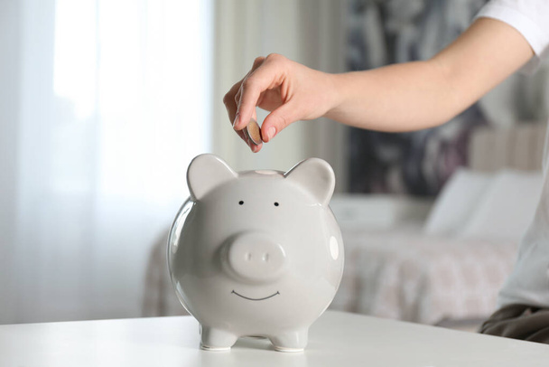 Woman putting money into piggy bank at white table indoors, closeup - Foto, Imagen