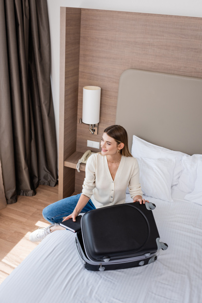 korkea kulma näkymä nuori nainen istuu sängyllä lähellä matkatavaroita ja tilalla muistikirja hotellihuoneessa  - Valokuva, kuva