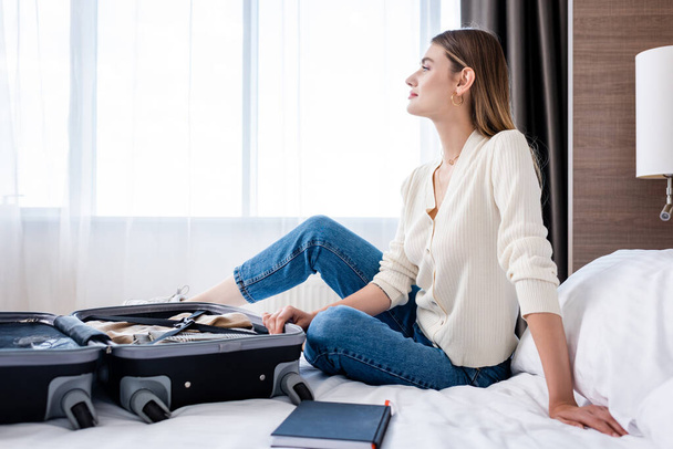 side view of young woman sitting on bed and looking away near luggage in hotel room  - Foto, afbeelding