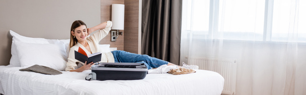 ホテルのベッドで荷物の近くのノートを見ている若い女性のパノラマの概念 - 写真・画像