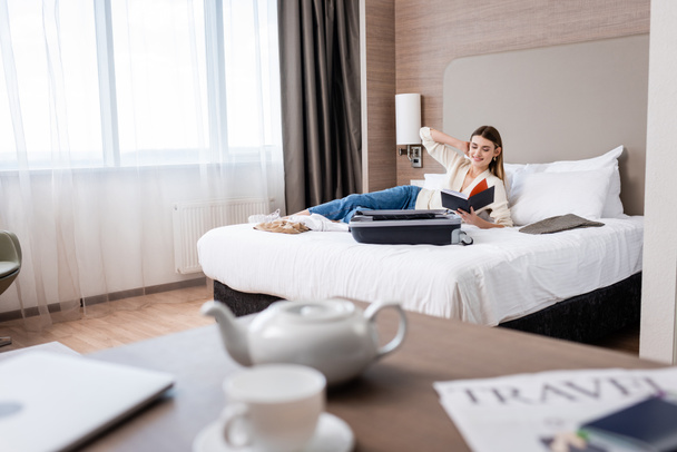 selective focus of young woman looking at notebook near luggage on bed in hotel - Photo, Image