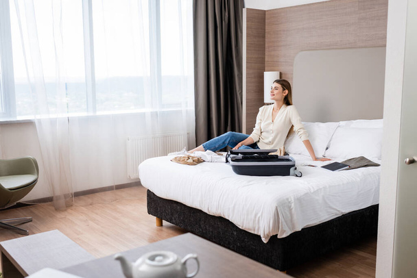 επιλεκτική εστίαση της νεαρής γυναίκας κοιτάζοντας μακριά κοντά σημειωματάριο και αποσκευές στο κρεβάτι στο ξενοδοχείο - Φωτογραφία, εικόνα