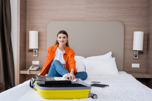young woman sitting on bed near yellow baggage and notebook in hotel  - 写真・画像