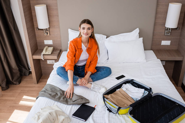 young woman sitting on bed near clothing, yellow baggage, smartphone and notebook in hotel  - 写真・画像