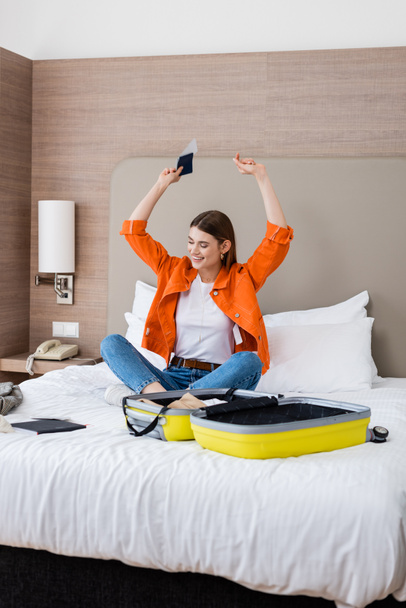 vzrušená žena držící pas a letenku nad hlavou v blízkosti zavazadel a notebooku na posteli v hotelu  - Fotografie, Obrázek