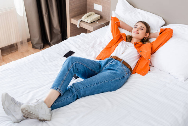 młoda kobieta odpoczywa w pobliżu smartfona z pustym ekranem na łóżku w hotelu - Zdjęcie, obraz