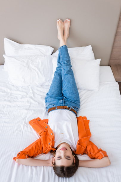vista de ángulo alto de mujer joven descalza en jeans descansando en la cama en la habitación de hotel - Foto, imagen
