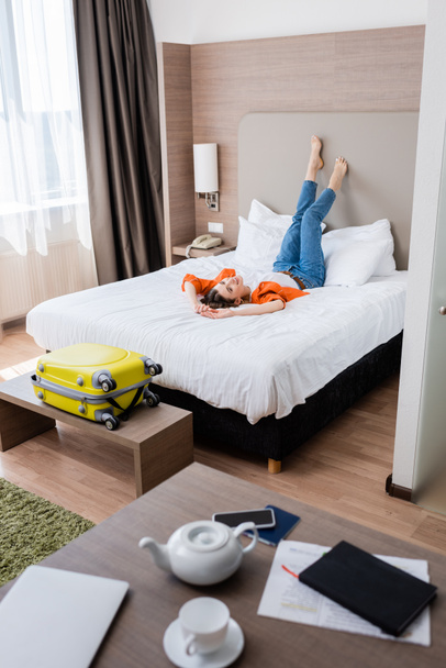 επιλεκτική εστίαση της ξυπόλυτη νεαρή γυναίκα στηρίζεται στο κρεβάτι στο σύγχρονο δωμάτιο του ξενοδοχείου - Φωτογραφία, εικόνα