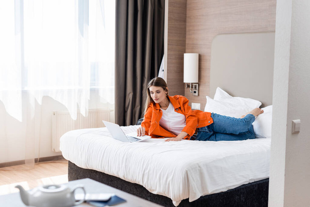 επιλεκτική εστίαση του ελεύθερου επαγγελματία ξαπλωμένος στο κρεβάτι και χρησιμοποιώντας φορητό υπολογιστή στο δωμάτιο του ξενοδοχείου - Φωτογραφία, εικόνα