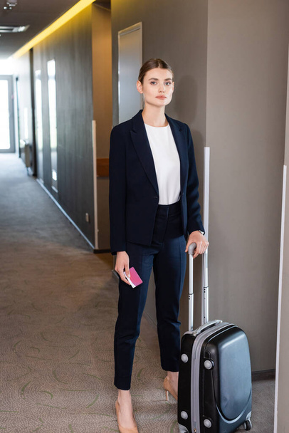 businesswoman in suit holding room card while standing with luggage in hall of hotel  - Φωτογραφία, εικόνα