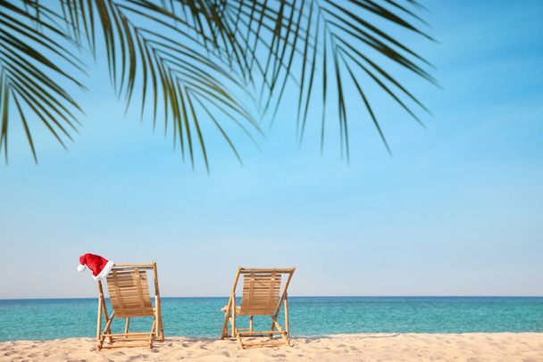 Sun loungers and Santa's hat on beach, space for text. Christmas vacation - Photo, Image