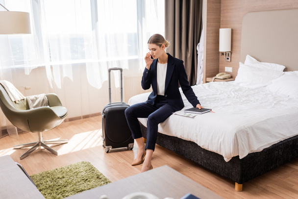 selective focus of businesswoman in suit talking on smartphone while sitting on bed near luggage in hotel  - Fotoğraf, Görsel