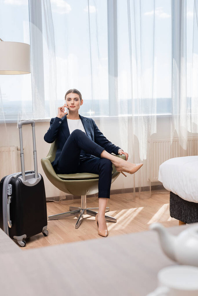 νεαρή επιχειρηματίας με κοστούμι μιλάει στο smartphone, ενώ κάθεται στην πολυθρόνα κοντά σε αποσκευές στο ξενοδοχείο  - Φωτογραφία, εικόνα
