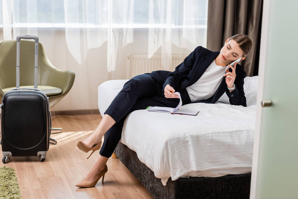 businesswoman in suit talking on smartphone and looking at notebook in hotel room  - Фото, изображение