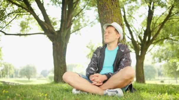 Mladý pohledný muž v klobouku sedí pod stromem v městském parku, pohyb kamery - Záběry, video