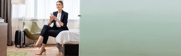 οριζόντια εικόνα της νεαρής επιχειρηματία με κοστούμι κρατώντας smartphone, ενώ κάθεται στο κρεβάτι κοντά σε ταξιδιωτική τσάντα στο δωμάτιο του ξενοδοχείου  - Φωτογραφία, εικόνα