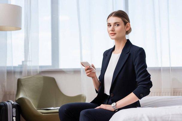 Geschäftsfrau im Anzug hält Smartphone in der Hand und blickt in die Kamera, während sie im Hotelzimmer auf dem Bett sitzt  - Foto, Bild