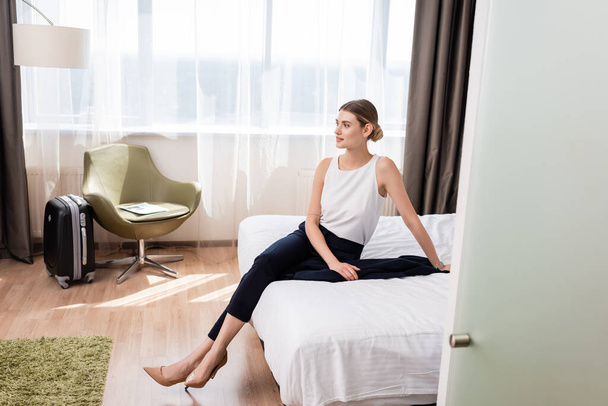 podnikatelka odvrací pohled, zatímco sedí na posteli v moderním hotelovém pokoji - Fotografie, Obrázek