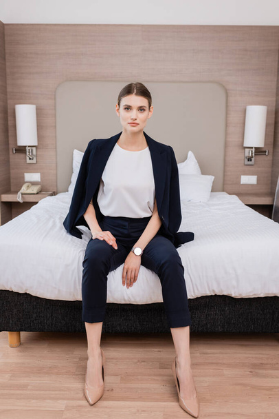 young businesswoman in suit looking at camera and sitting on bed in hotel  - 写真・画像